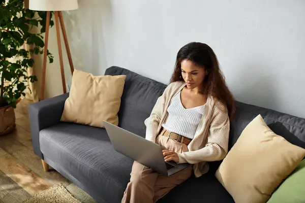 Una giovane donna si concentra intensamente sul suo computer portatile mentre seduto comodamente nel suo spazio di vita chic. — Foto stock