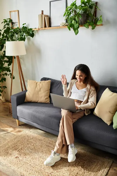 Une femme s'assoit confortablement sur un canapé, interagissant joyeusement sur son ordinateur portable dans un espace lumineux. — Photo de stock