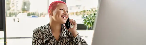 A bald woman with a bright pink haircut smiles as she talks on her phone at her workspace. — Stock Photo