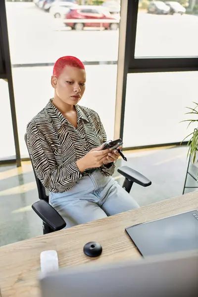 Eine glatzköpfige Frau mit leuchtend roten Haaren nutzt ihr Handy, während sie an einem stilvollen Schreibtisch sitzt. — Stockfoto