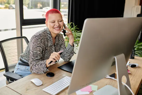 Cheerful bald woman talks on the phone, smiling while working on her laptop in a creative space. — Stock Photo