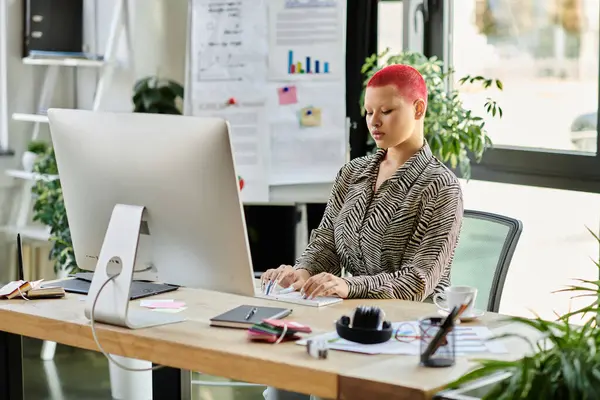 A bald woman with vibrant hair color focuses on work at a sleek desk amidst greenery. — Stock Photo