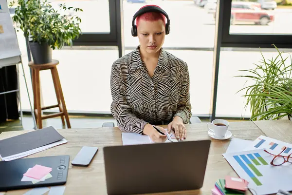A dedicated bald woman takes notes while engaged with her laptop in a well lit workspace. — Stock Photo