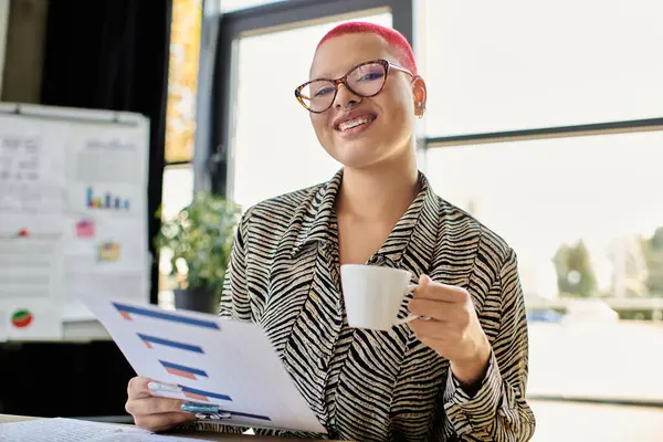 The bald woman in a stylish outfit smiles as she holds a cup, engaging with her work materials. — Stock Photo