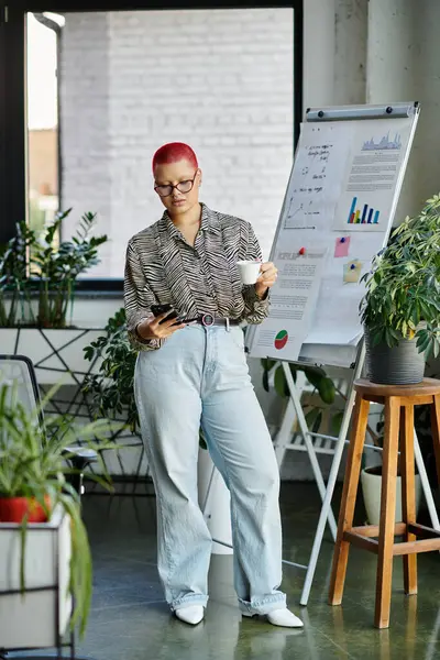 A confident bald woman sips coffee and checks her phone amidst lush office greenery — Stock Photo
