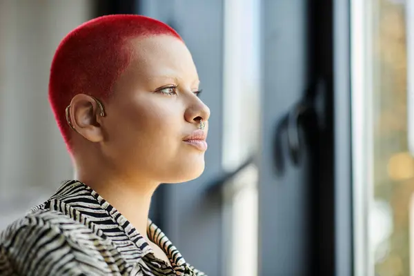 A bald woman with striking red hair reflects quietly while looking outside in a warm atmosphere. — Stock Photo