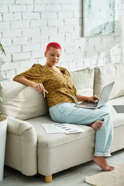 A bold and stylish woman sits comfortably on a couch, focused on her laptop while working. — Stock Photo
