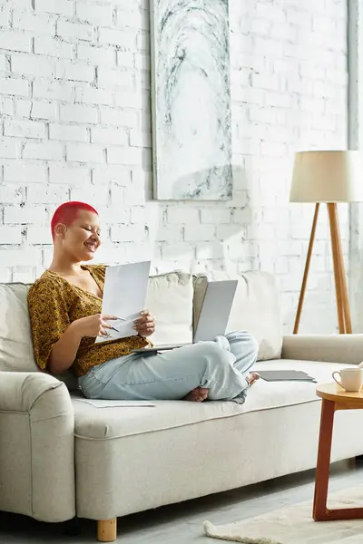 A bald woman sitting comfortably on a couch, smiling as she works on her laptop. — Stock Photo