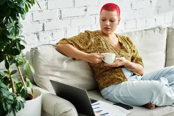 A bald woman with striking hair color sips tea while relaxing on her couch, immersed in thought. — Stock Photo