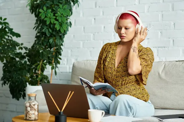 A bald woman sits comfortably on a couch, lost in her book while wearing headphones. — Stock Photo