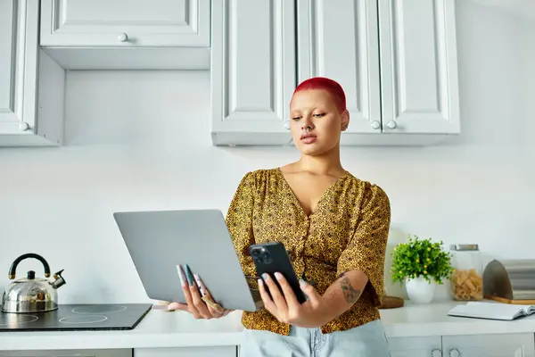 This beautiful bald woman engages with her laptop and smartphone in a sunny kitchen. — Stock Photo