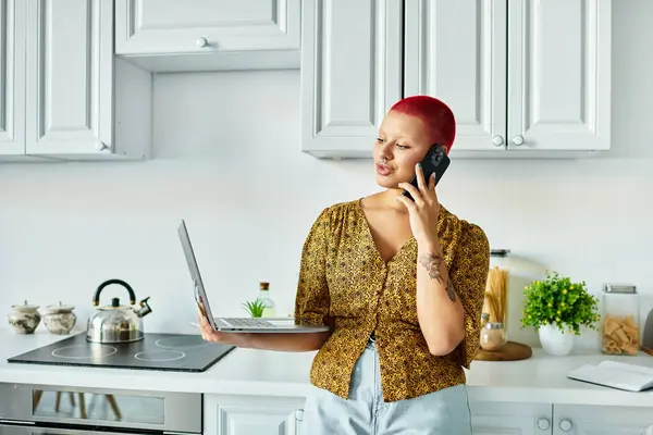 Una mujer calva audaz se involucra en una llamada, equilibrando a la perfección su computadora portátil en un entorno de cocina moderna. - foto de stock