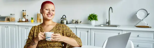 A cheerful bald woman sips tea while engaged with her laptop in a bright kitchen. — Stock Photo