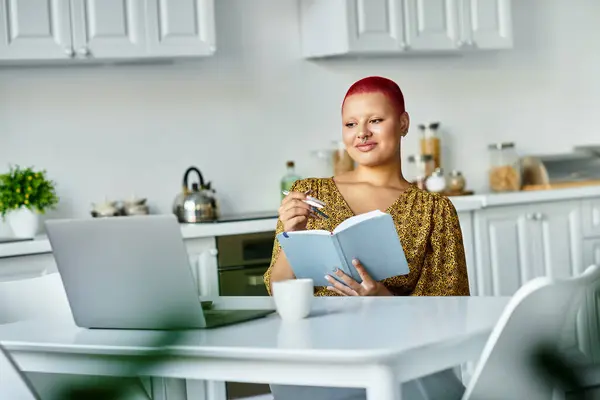 A bald woman sits at a table in a bright kitchen, reflecting while journaling with a drink nearby. — Stock Photo