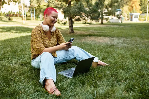 A bald woman with vibrant hair relaxes on the grass, engaging with her devices and smiling. — Stock Photo