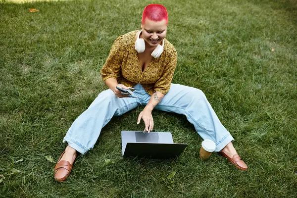 Una donna calva gioiosa con i capelli colorati gode di lavorare sul suo computer portatile mentre seduto sull'erba. — Foto stock