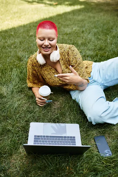 A cheerful bald woman sits on the grass, sipping coffee and engaging with her laptop. — Stock Photo