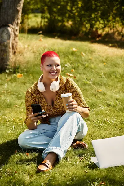A joyful bald woman relaxes in nature, sipping coffee and using her phone, surrounded by greenery. — Stock Photo