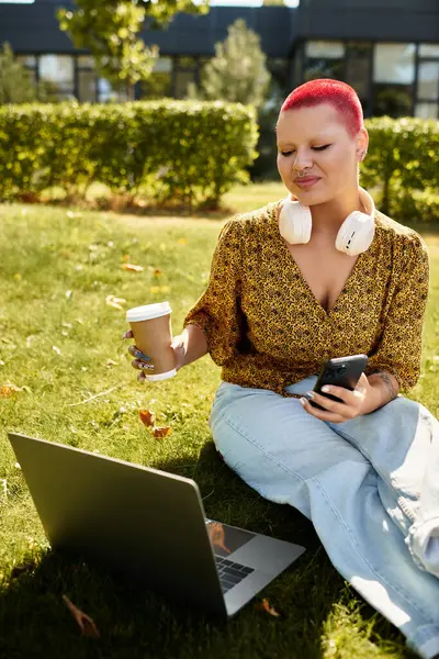 A happy bald woman enjoys a warm drink and engages with her laptop while sitting on grass. — Stock Photo
