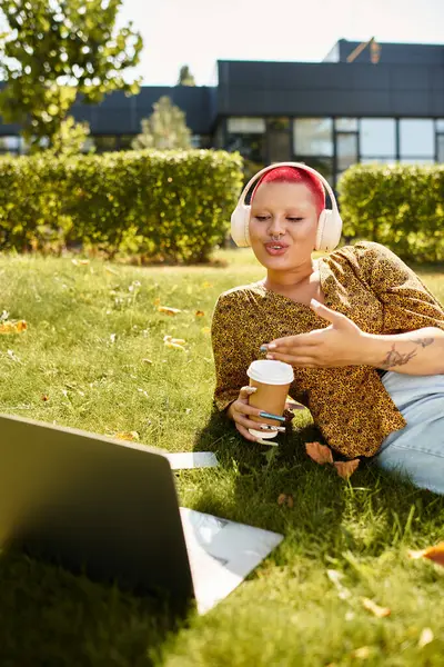 A cheerful bald woman relaxes on the grass, sipping coffee while engaged in her laptop work. — Stock Photo