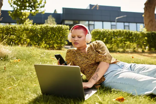 A bald woman wearing headphones engages with her laptop and phone while lounging outdoors. — Stock Photo