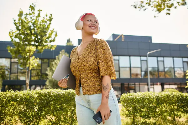 A cheerful bald woman walks outside, enjoying the warm sun while holding her laptop and smiling. — Stock Photo