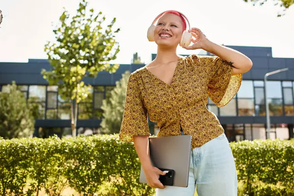 A joyful bald woman listens to music with headphones, holding her laptop while smiling outside. — Stock Photo
