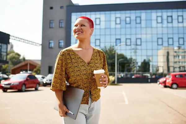A bald woman with vibrant hair color walks happily, holding coffee and a laptop. — Stock Photo