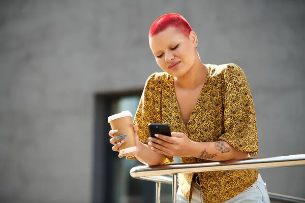 A bald woman with vibrant red hair drinks coffee while checking her smartphone outdoors. — Stock Photo