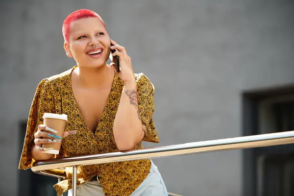 Una mujer calva radiante sonríe mientras sostiene el café y habla por teléfono al aire libre. - foto de stock