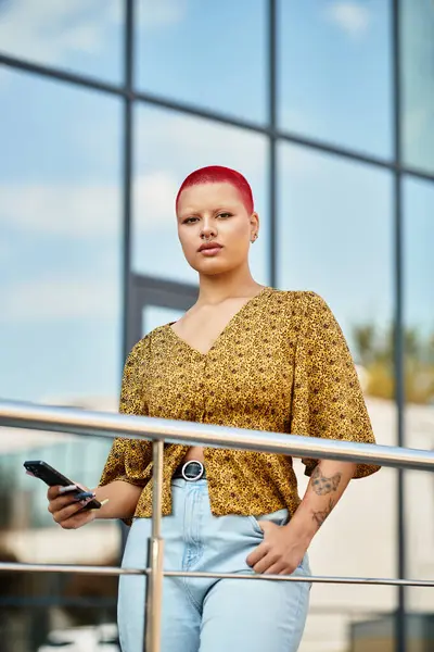 The beautiful bald woman poses confidently on a balcony, holding a phone against a bright sky. — Stock Photo