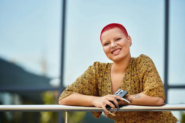 A radiant bald woman, dressed in a stylish outfit, smiles cheerfully while holding a small device. — Stock Photo