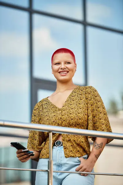 Una donna calva con i capelli rossi sorride mentre tiene un telefono su un balcone soleggiato. — Foto stock