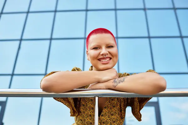 A radiant bald woman with bright red hair smiles while leaning on a railing under clear blue skies. — Stock Photo