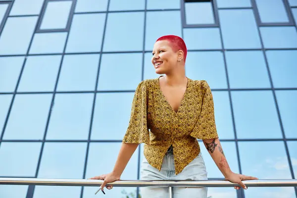 Una mujer calva vibrante sonríe brillantemente mientras se apoya en una barandilla de balcón contra un fondo llamativo. - foto de stock