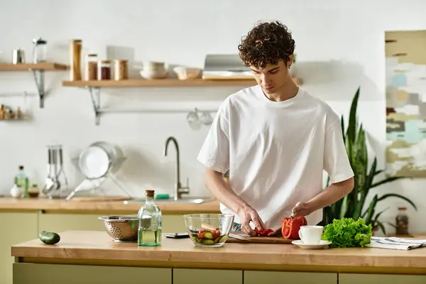 Dans une cuisine élégante, un beau jeune homme aux cheveux bouclés côtelent habilement les légumes pour une salade vibrante à déguster. — Photo de stock