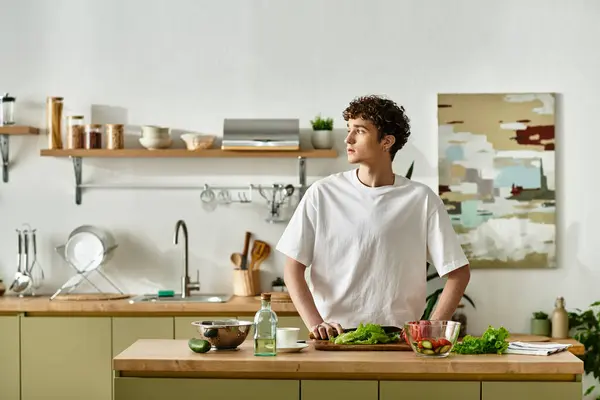 In a modern kitchen, a young man with curly hair thoughtfully prepares a vibrant salad, embracing healthy living. — Stock Photo
