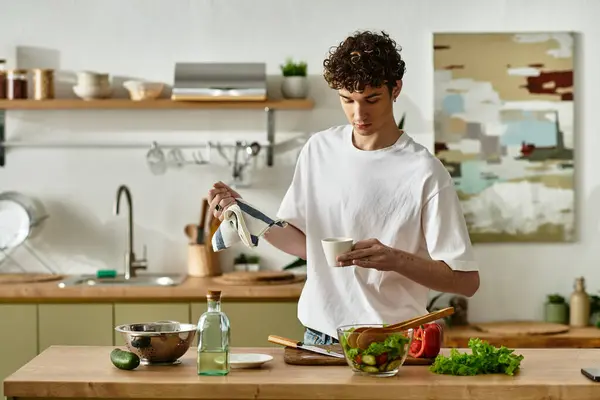 Ein hübscher junger Mann mit lockigem Haar trinkt Kaffee in der Nähe von frischem Salat in einer stilvollen, modernen Küche. — Stock Photo