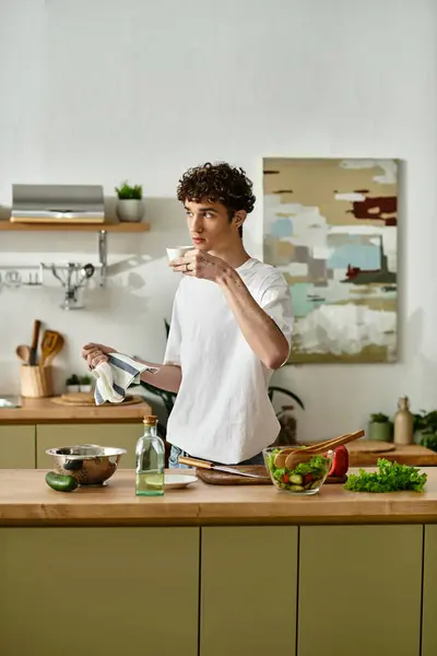 In a sleek kitchen, a handsome young man with curly hair drinking coffee near fresh salad — Stock Photo