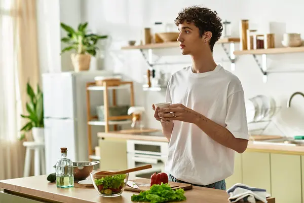 A handsome young man with curly hair enjoys making a healthy salad in his stylish kitchen filled with plants. — Stock Photo