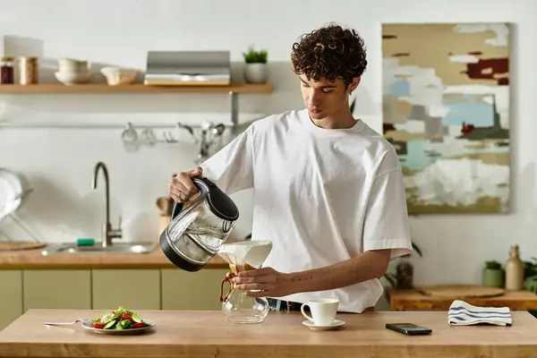 En una cocina contemporánea, un joven guapo vierte café de una tetera en un vaso, disfrutando de momentos culinarios. - foto de stock