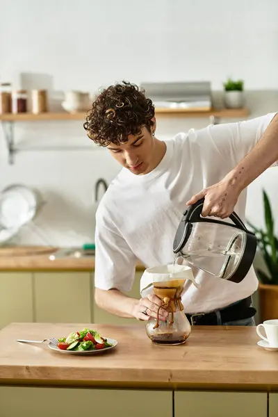 Un joven guapo vierte café caliente en una olla de cerveza mientras organiza una ensalada en una cocina luminosa y acogedora.. - foto de stock