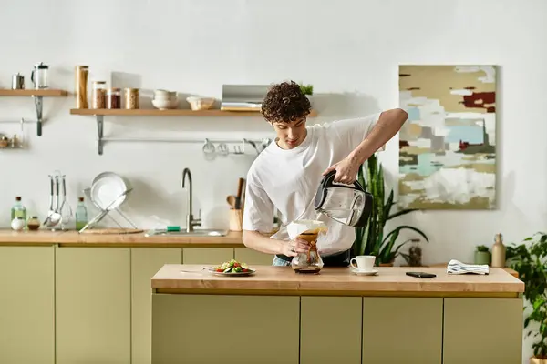 En una cocina luminosa, un joven apuesto vierte café en una taza, saboreando un tranquilo descanso matutino. - foto de stock