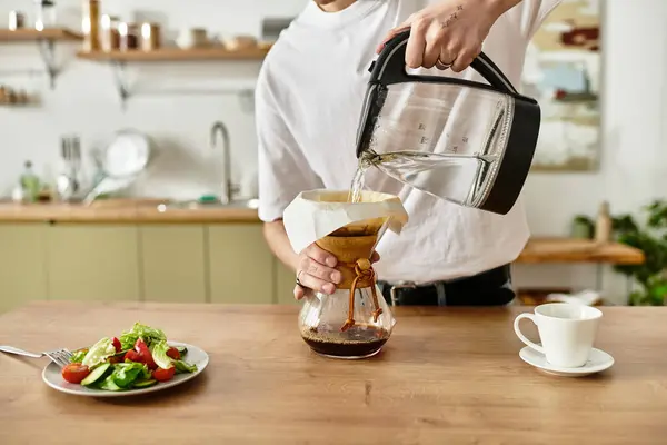 En una acogedora cocina, un hombre vierte agua caliente sobre el café molido mientras una ensalada espera en la mesa. - foto de stock