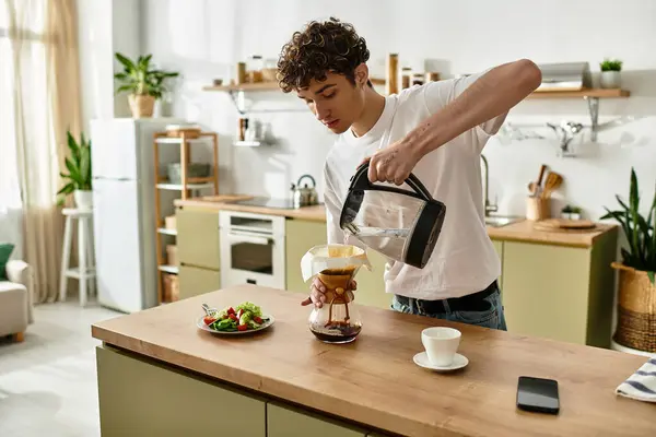 Un joven guapo vierte cuidadosamente agua caliente para la preparación del café, rodeado de una ensalada fresca y una decoración acogedora. - foto de stock