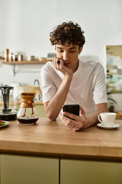 In a warm kitchen, a handsome young man with curly hair savors his coffee while focusing intently on his phone. — Stock Photo