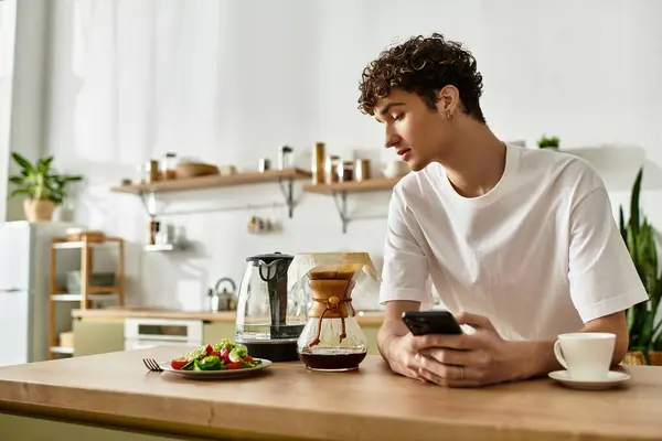 A handsome young man with curly hair sits at a table, sipping coffee while checking his phone, surrounded by food. — Stock Photo