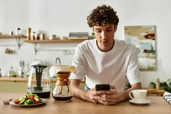 A handsome young man with curly hair is savoring a warm cup of coffee while checking his phone in a bright kitchen. — Stock Photo