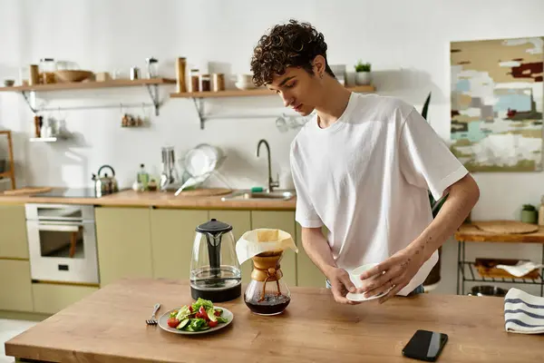 Ein junger Mann mit lockigem Haar brüht Kaffee und bereitet in einer hellen, einladenden Küche einen frischen Salat zu.. — Stockfoto