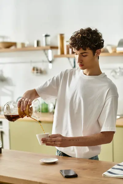 Un joven guapo con el pelo rizado vierte café recién hecho en una taza, disfrutando de un momento en su cocina. - foto de stock